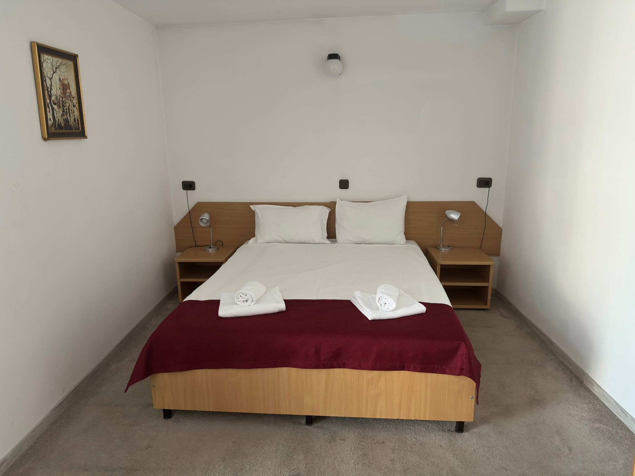 Un lit bien fait, avec une couverture rouge, deux oreillers blancs et deux serviettes enroulées. Il y a deux tables de chevet avec des lampes, une applique murale et un tableau encadré accroché au mur.