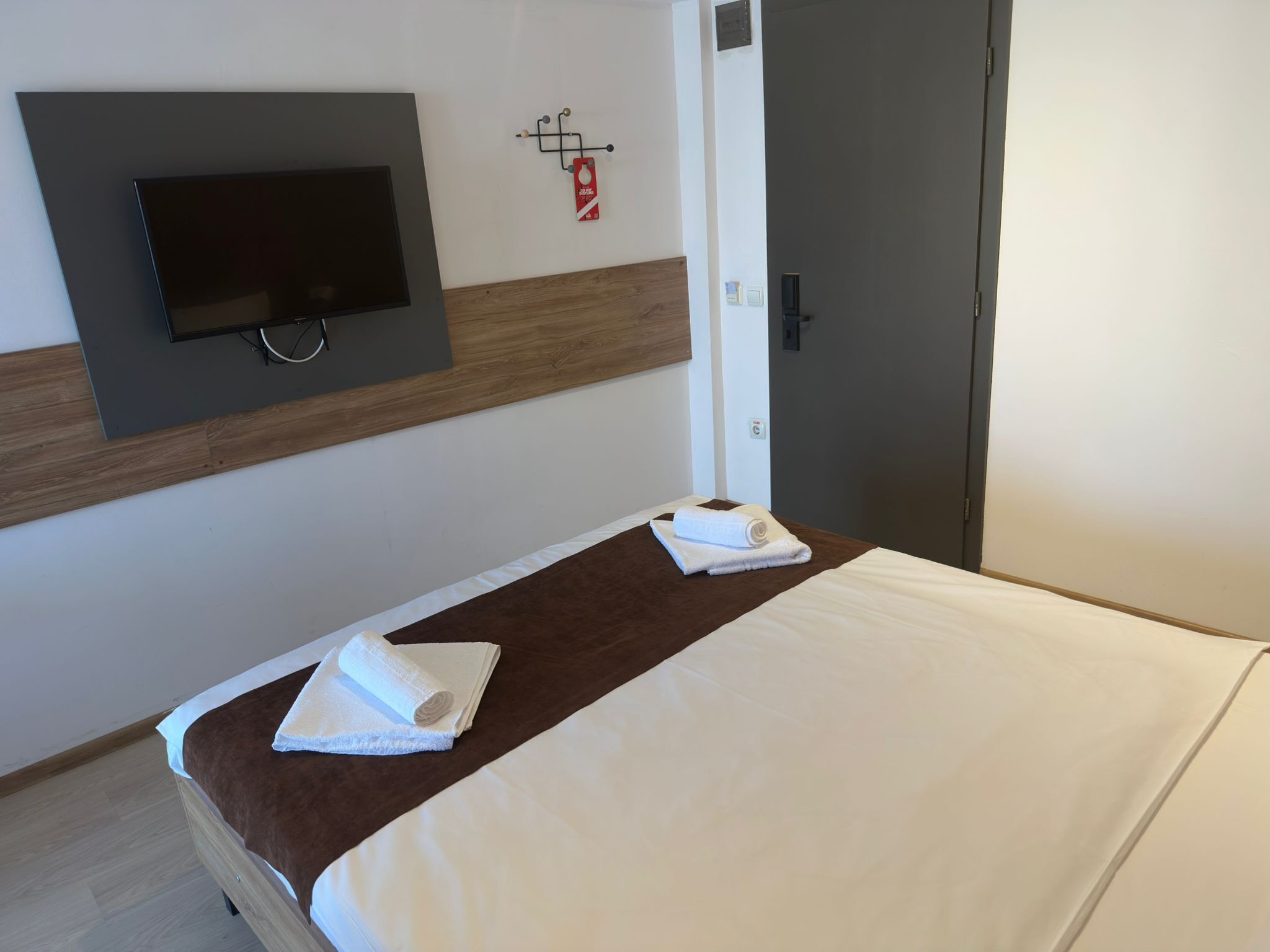 A hotel room with a bed, a TV mounted on the wall, a wooden panel, and a door.