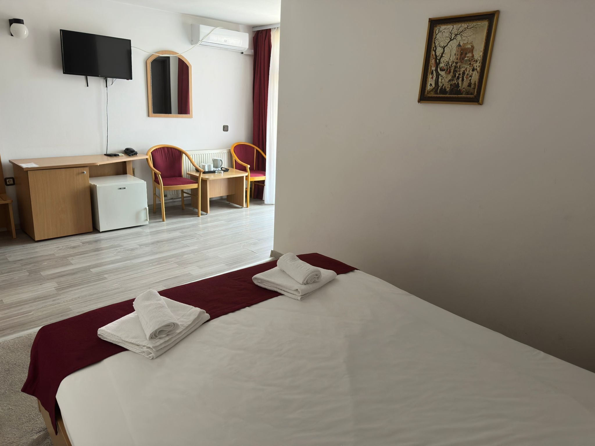 A hotel room with a bed, a desk, two chairs, a small fridge, a TV, and a painting on the wall.