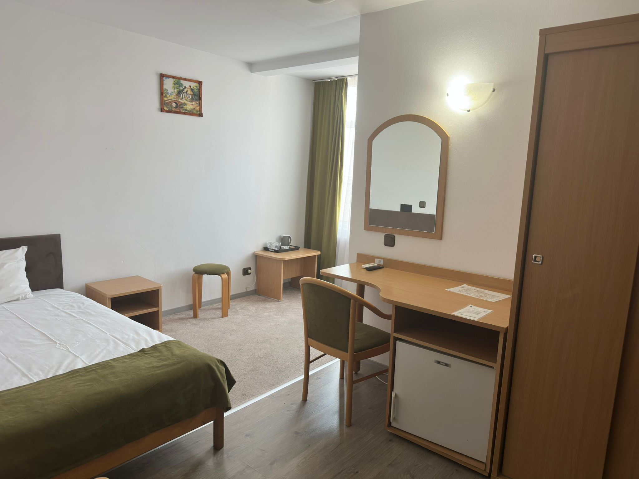 Une chambre d’hôtel avec un lit, un bureau, une chaise, une petite armoire, un miroir et un tableau accroché au mur.