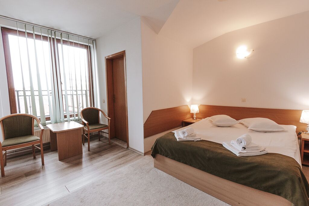 Une chambre d’hôtel avec un lit, deux chaises, une petite table et une fenêtre avec des stores.