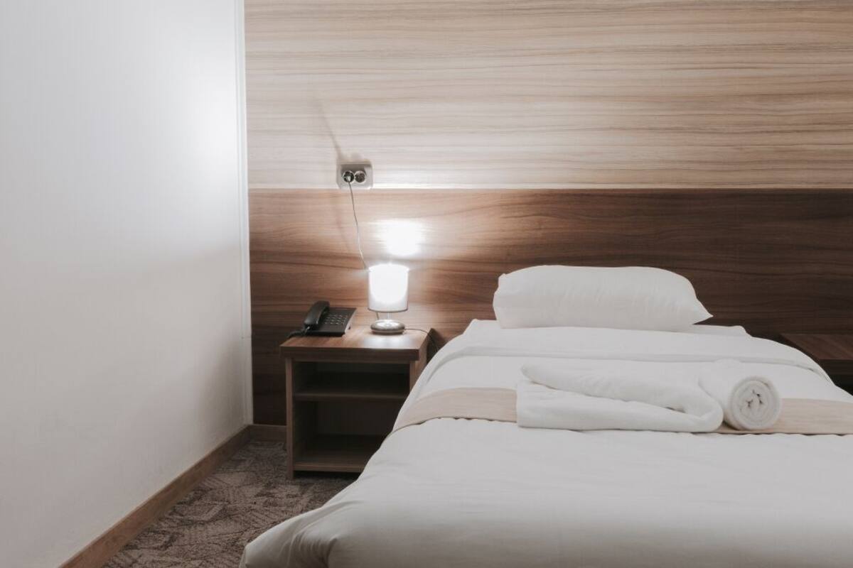 Quarto de hotel com cabeceira de madeira, mesa de cabeceira com abajur, telefone e toalhas dobradas com cuidado.