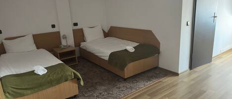 Habitación de hotel con dos camas individuales, una mesita de noche pequeña con lámpara, una alfombra estampada y una puerta a la derecha.