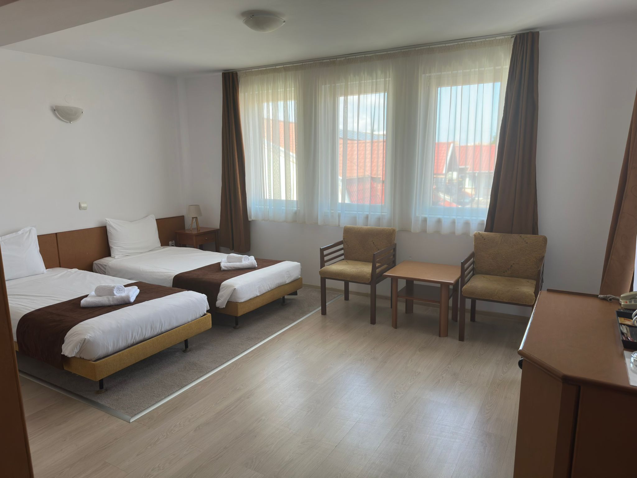 A hotel room with two single beds, a small table, two armchairs, and a desk with a telephone.