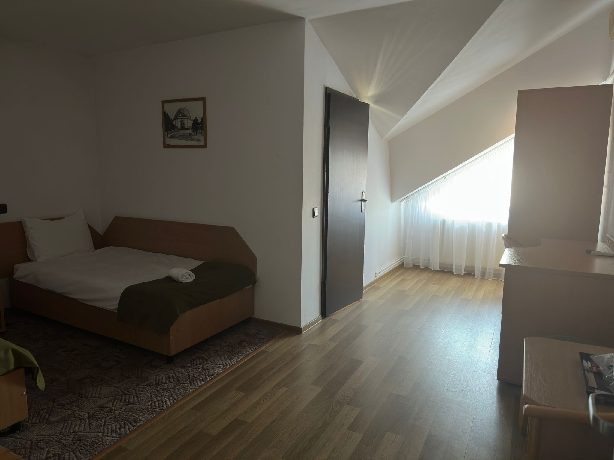 A hotel room with a bed, a desk, a chair, a door, and a window with curtains.