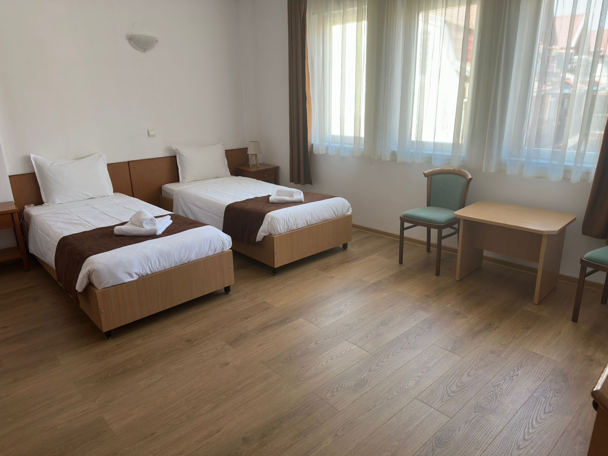 Een hotelkamer met twee éénpersoonsbedden, een bureau, een stoel en een raam met dunne gordijnen.