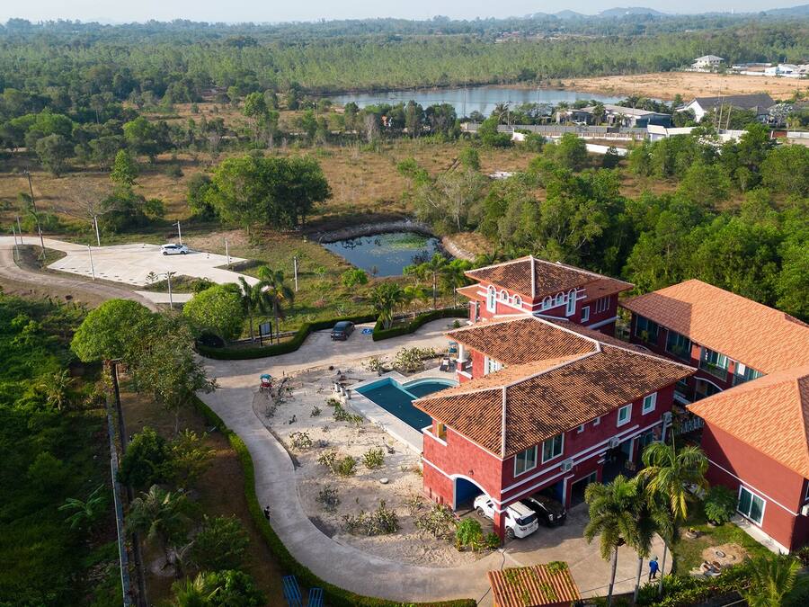 A large, multi-story building with a red roof and multiple windows, surrounded by lush greenery and a small pond.