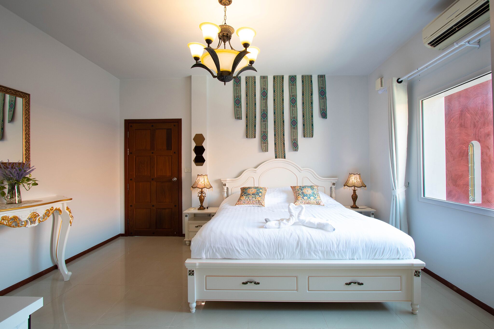 A neatly arranged bedroom with a large bed, bedside lamps, a chandelier, and a view of the outdoors.