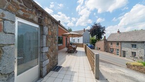 Ein Steinhaus mit einer gefliesten Terrasse, einer Glasdoor und einem Tisch mit Stühlen unter einem Sonnensegel.