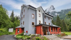 Edifici de dues plantes amb façana gris i accents vermells, envoltat de verdura abundant i muntanyes.