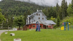 Una casa amb façana vermella i gris, un parc infantil amb tobogan i espigues, i un paisatge de muntanyes boscoses al fons.