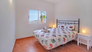 A bedroom with a bed, a nightstand, a window with a view of palm trees, and a tiled floor.