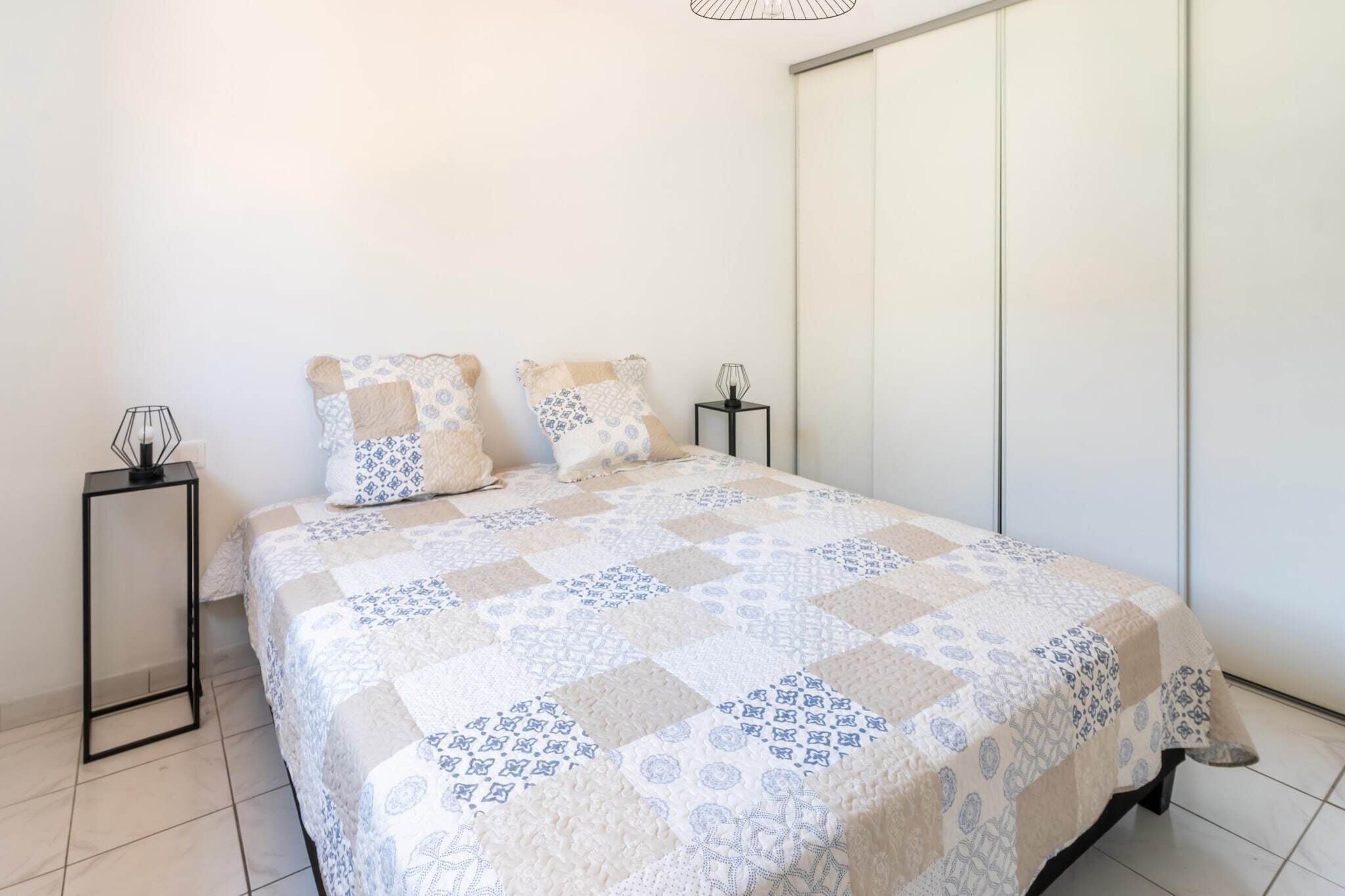 Uma cama bem arrumada com um edredom estampado, uma mesinha de cabeceira com abajur e um guarda-roupa na parede.
