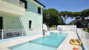 A rectangular swimming pool with a water fountain, surrounded by a white fence. - Belvilla by OYO Appartamento ROSSO (Jesolo)