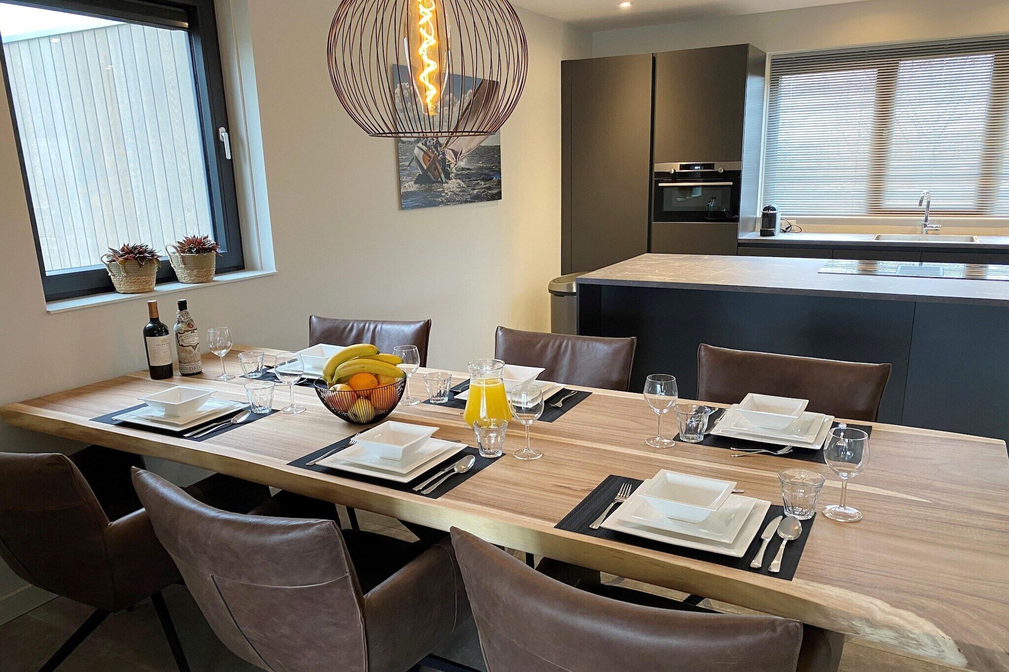 A dining area with a long wooden table set for six, surrounded by leather chairs. There is a small island with a sink and a refrigerator in the background.