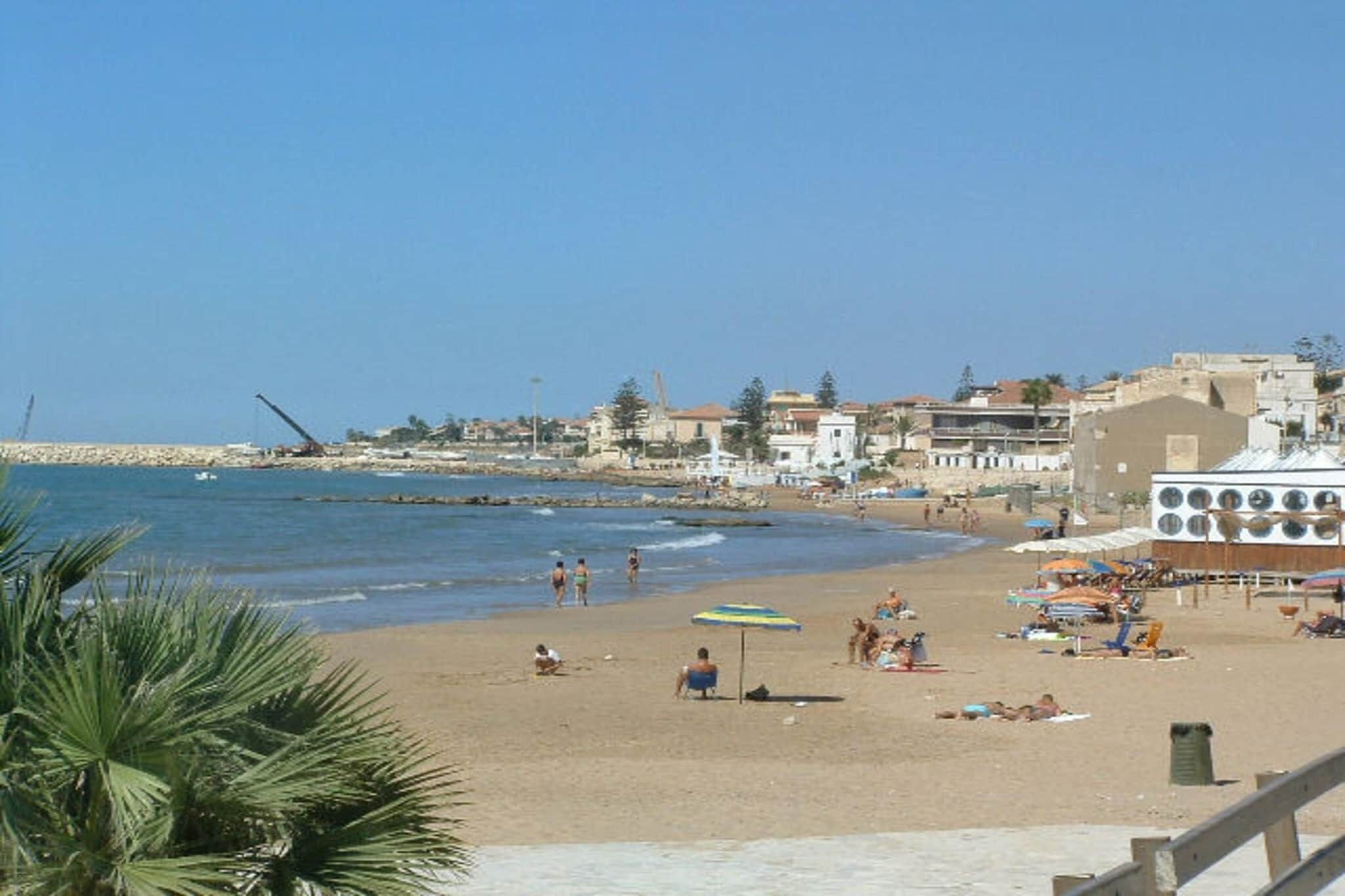 Ein Strand mit badenden und sonnenden Menschen, einigen Sonnenschirmen und Gebäuden im Hintergrund.