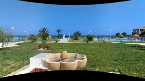 Een aangelegde tuin met een centraal waterpartij, palmbomen en een helder uitzicht op de zee.
