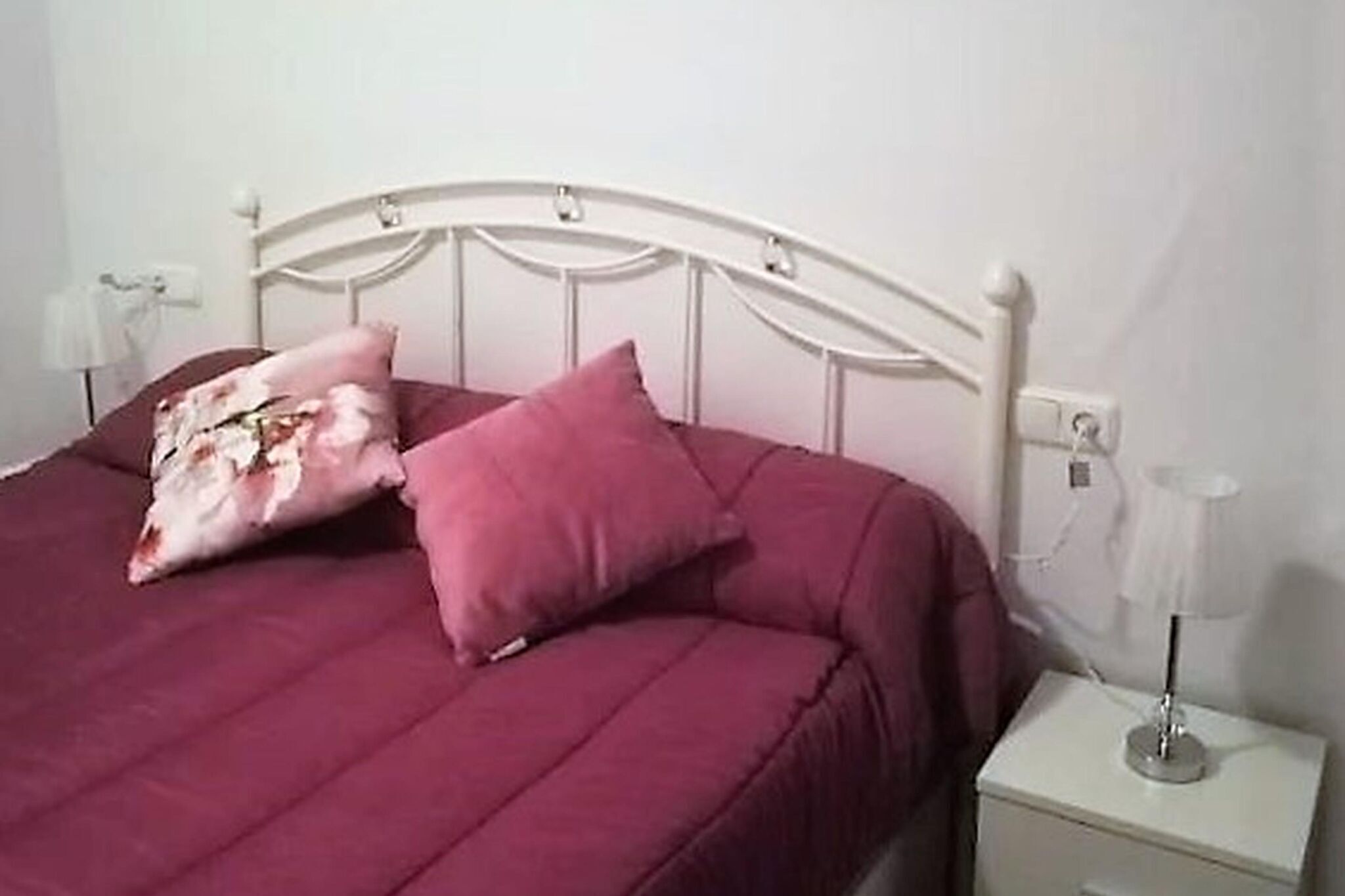 Un lit avec une tête de lit en métal blanche, une couvre-lit rose et deux oreillers. Il y a une table de chevet blanche avec une lampe et une prise de courant.