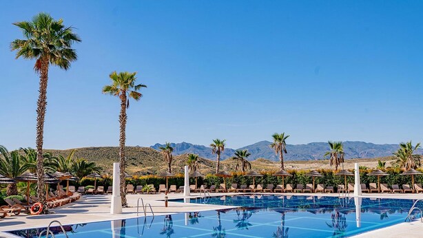 Zona de piscina con sillones de descanso y sombrillas, palmeras y montañas al fondo.