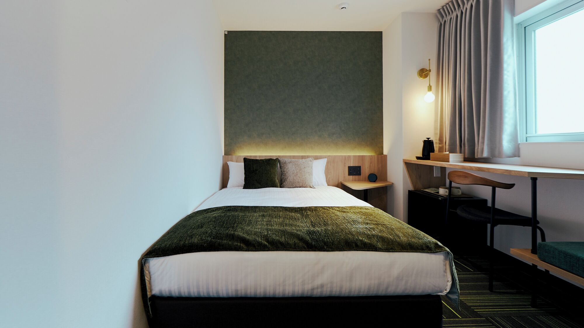 A hotel room with a bed, a desk, a chair, a nightstand, and a window with curtains.