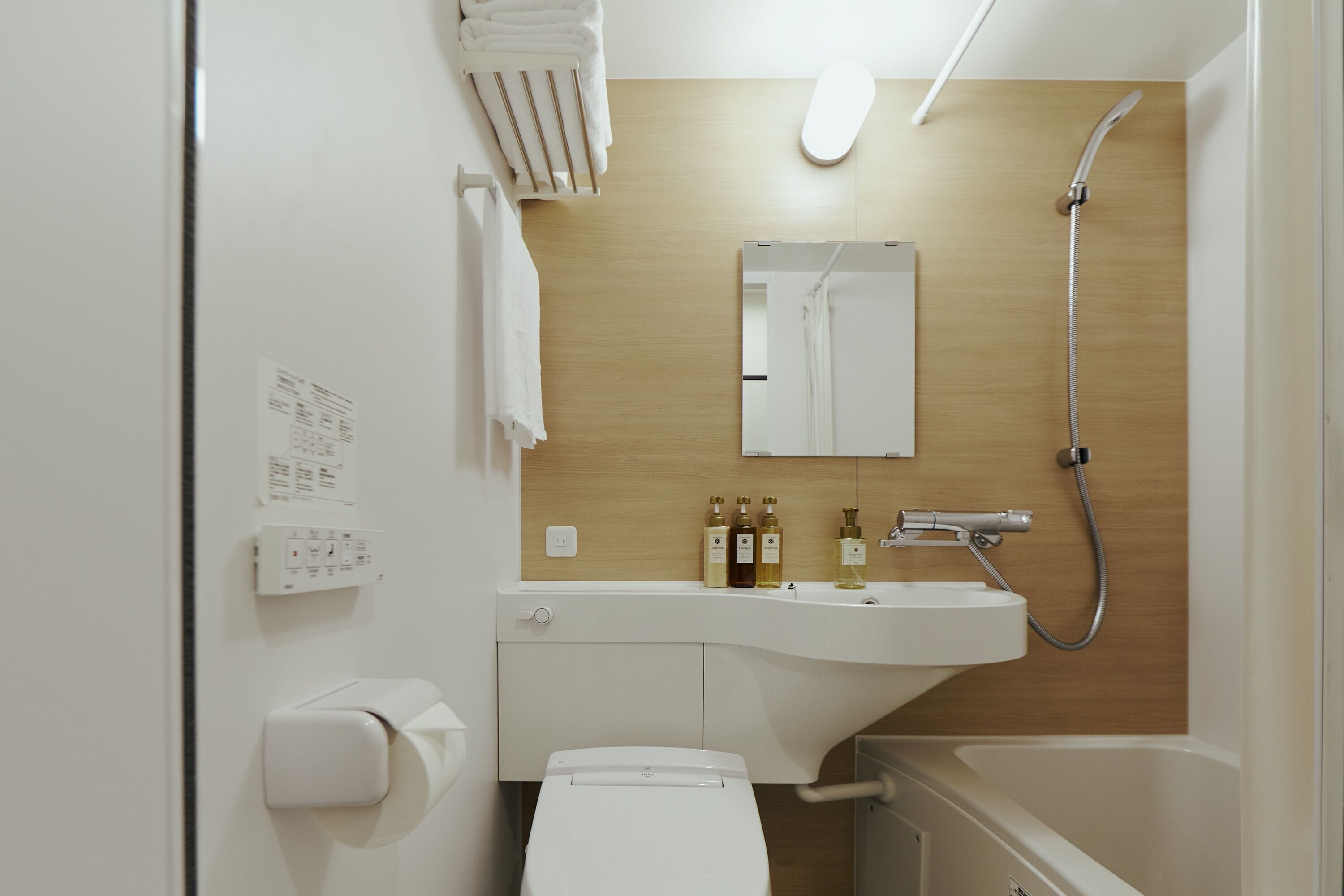 Une salle de bain moderne équipée de toilettes, d’un lavabo et d’une douche. On y trouve des serviettes suspendues à un porte-serviettes, un miroir au-dessus du lavabo et un panneau de commande fixé au mur.