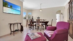 A living room with a dining area, a purple armchair, a table with magazines, and a television displaying a beach scene.