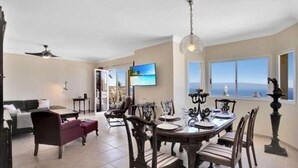 A spacious dining area with a large table and chairs, a television mounted on the wall, and a view of the ocean through large windows.