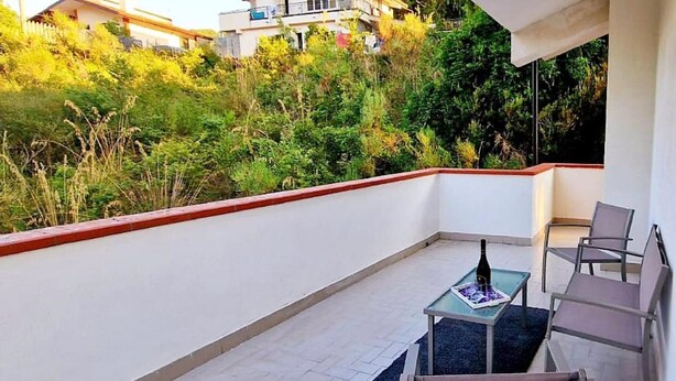 Un balcón con mesa y sillas, rodeado de vegetación y edificios al fondo.