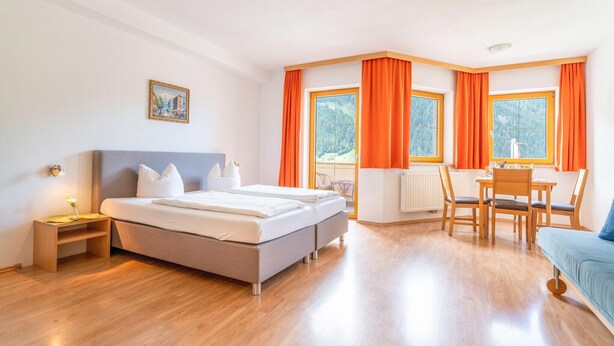 A modern hotel room with a large bed, a dining area, and large windows with orange curtains. - Apartment in Neustift near Ski Lift (Neustift im Stubaital)