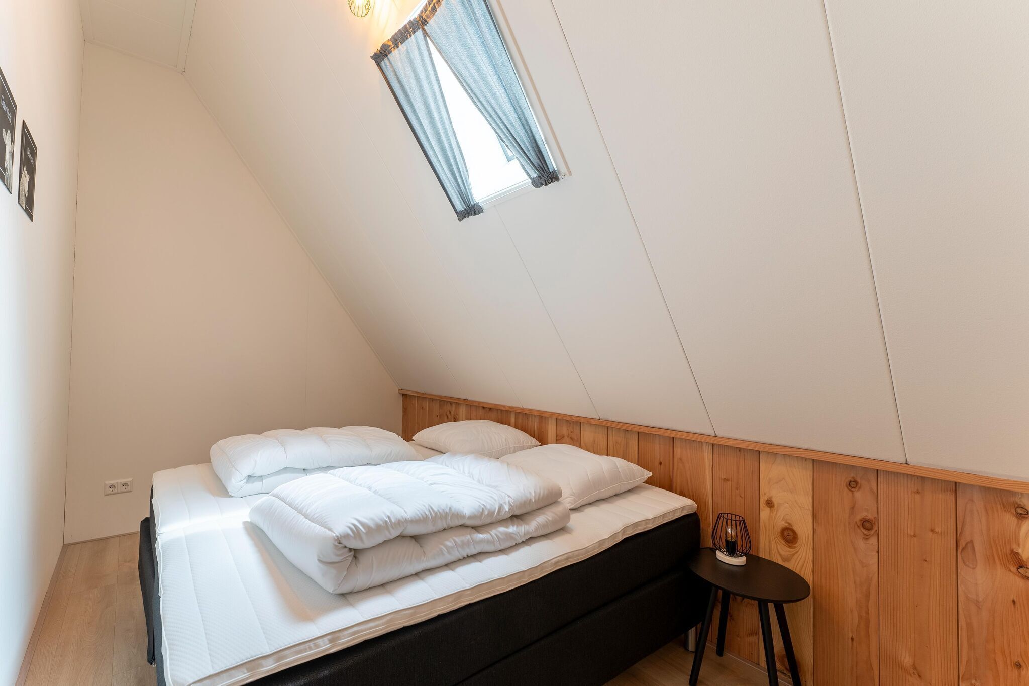 A bedroom with a bed, a small table, a window with curtains, and wooden paneling on the walls.