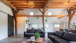Un salón amplio con techo alto, vigas de madera, un sofá negro, una mesa de centro redonda y plantas colgantes.