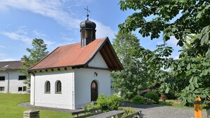 A small building with a red-tiled roof, a cross on top, and a garden with benches and a fountain. - Geraeumige Wohnung für 5 Personen in Drachselsried (Drachselsried)