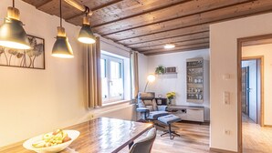 A dining area with a wooden table, chairs, and pendant lights. There is a window with curtains, a chair with a plant, and a shelf with glasses. - Geraeumige Wohnung für 5 Personen in Drachselsried (Drachselsried)