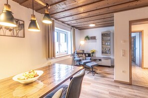 Un comedor con mesa de madera, sillas y lámparas colgantes. Se ve una ventana con cortinas, una silla con una planta y un estante con vasos.