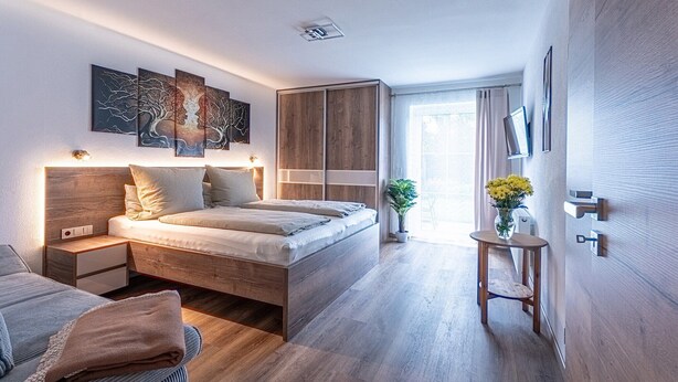 A modern bedroom with a wooden bed, bedside tables, a nightstand, a small table with a vase of flowers, and a sliding wardrobe. - Geraeumige Wohnung für 5 Personen in Drachselsried (Drachselsried)