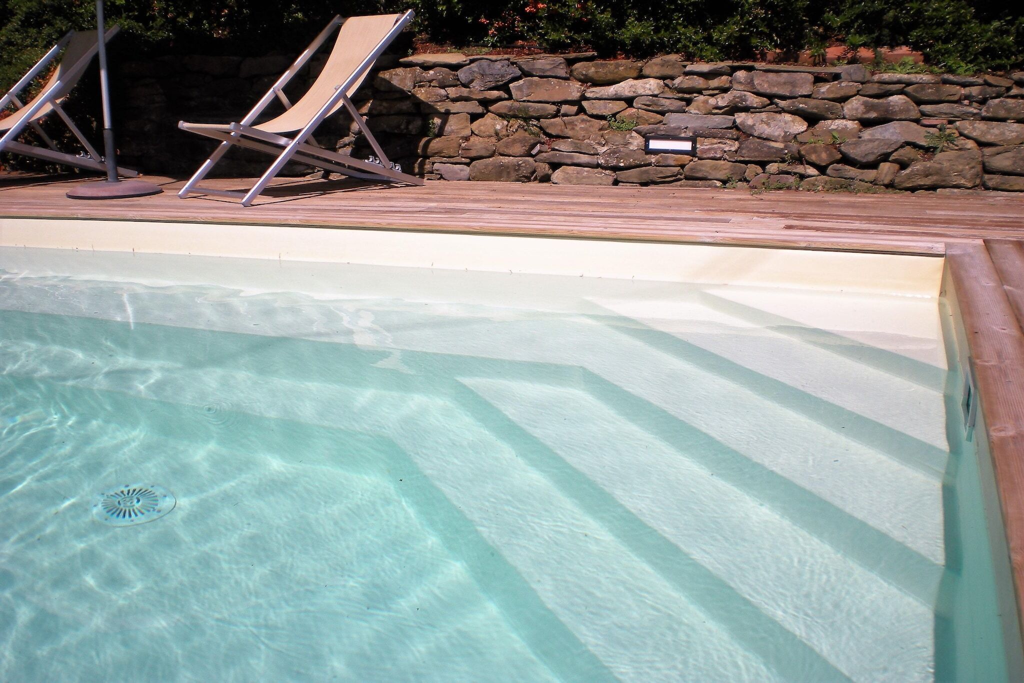 A swimming pool with clear water, a stone wall, and two lounge chairs.