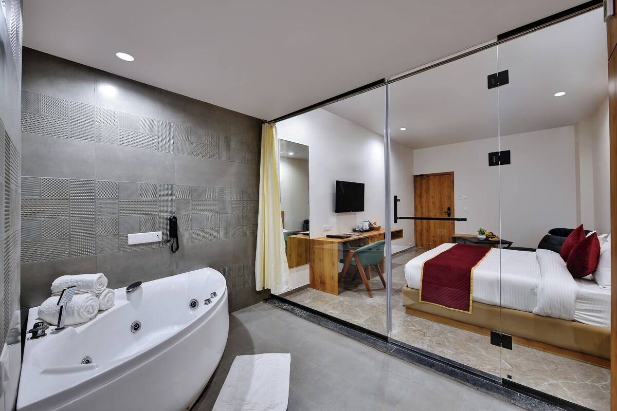 A modern hotel room with a large bathtub, a desk with a chair, a bed with red pillows, and a television on the wall.