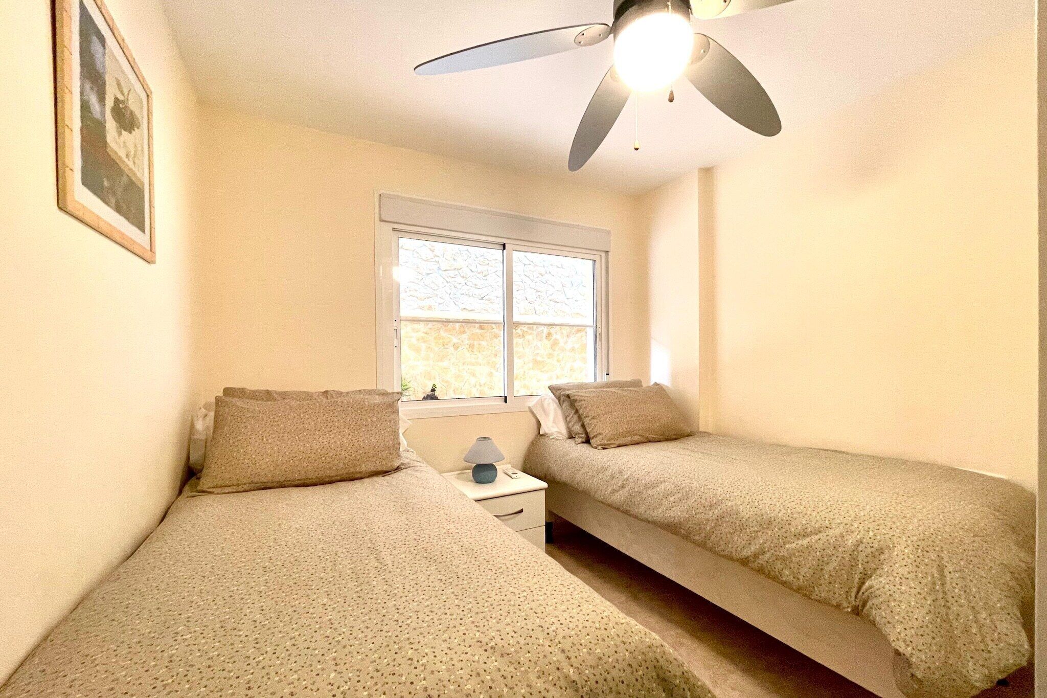 A bedroom with two single beds, a ceiling fan, a nightstand with a lamp, and a window with a view of the outside.