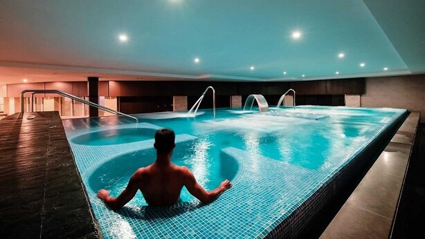 Una piscina interior con una persona sentada en el borde, rodeada de suelo y paredes con baldosas, y un techo con iluminación empotrada.