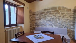 Ein gemütliches Zimmer mit Steinwand, einem Holztisch mit Unterlage, einem weißen Sofa und einem Fenster mit Holzläden.
