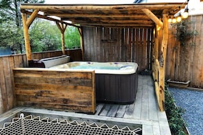 Una terraza de madera con un jacuzzi, una banca de madera y una pérgola con luces colgantes.