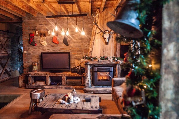 Un interior rústico con pared de piedra, techo de madera, chimenea, televisor y adornos navideños.