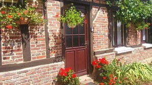 Een gebouw van baksteen met een donkerrode deur, bloembakken met bloemen en een kleine tuin met rode bloemen en groene planten.