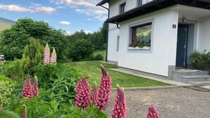A white building with a modern design, a visible entrance, and a well-maintained garden with pink flowers.