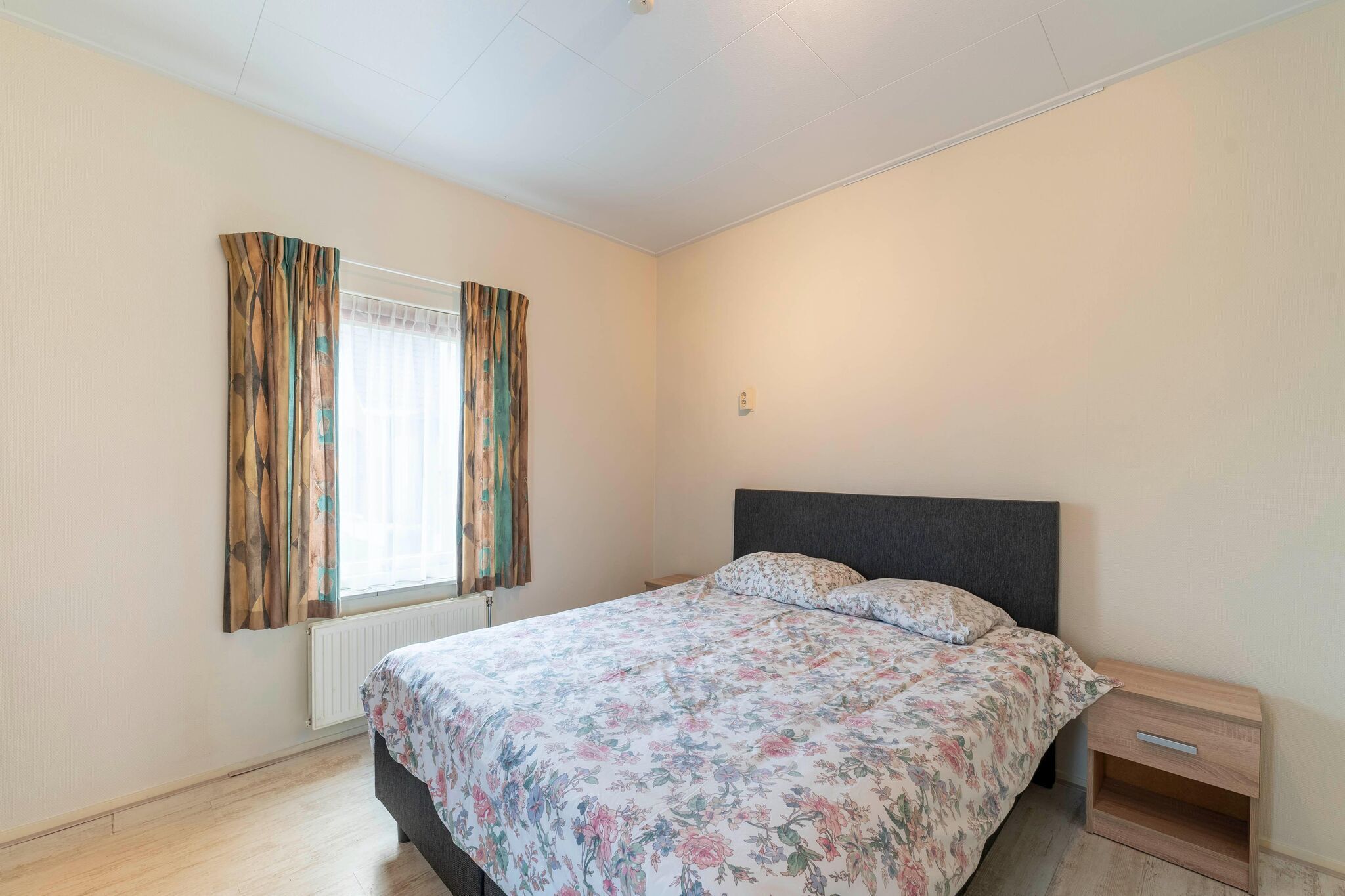 Un dormitorio con cama, mesita de noche, ventana con cortinas y radiador.