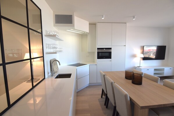 Una cocina moderna con armarios blancos, una zona de comedor con mesa y sillas de madera, y un televisor de pantalla plana montado en la pared.