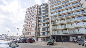 Un edificio residencial de varios pisos con balcones, una zona de estacionamiento y una tienda.