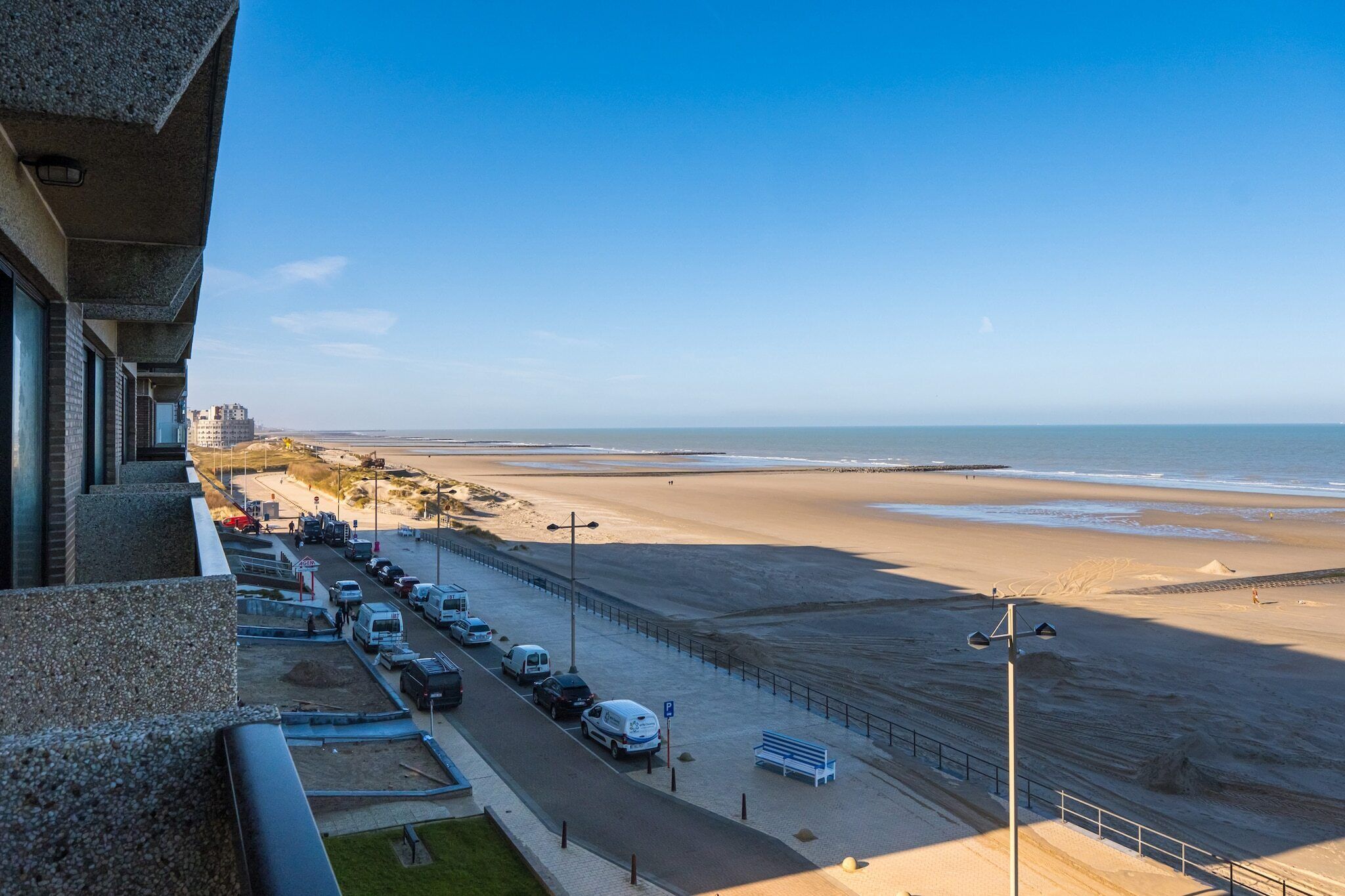A beachside parking area with vehicles, a pedestrian walkway, and a view of the beach and ocean.