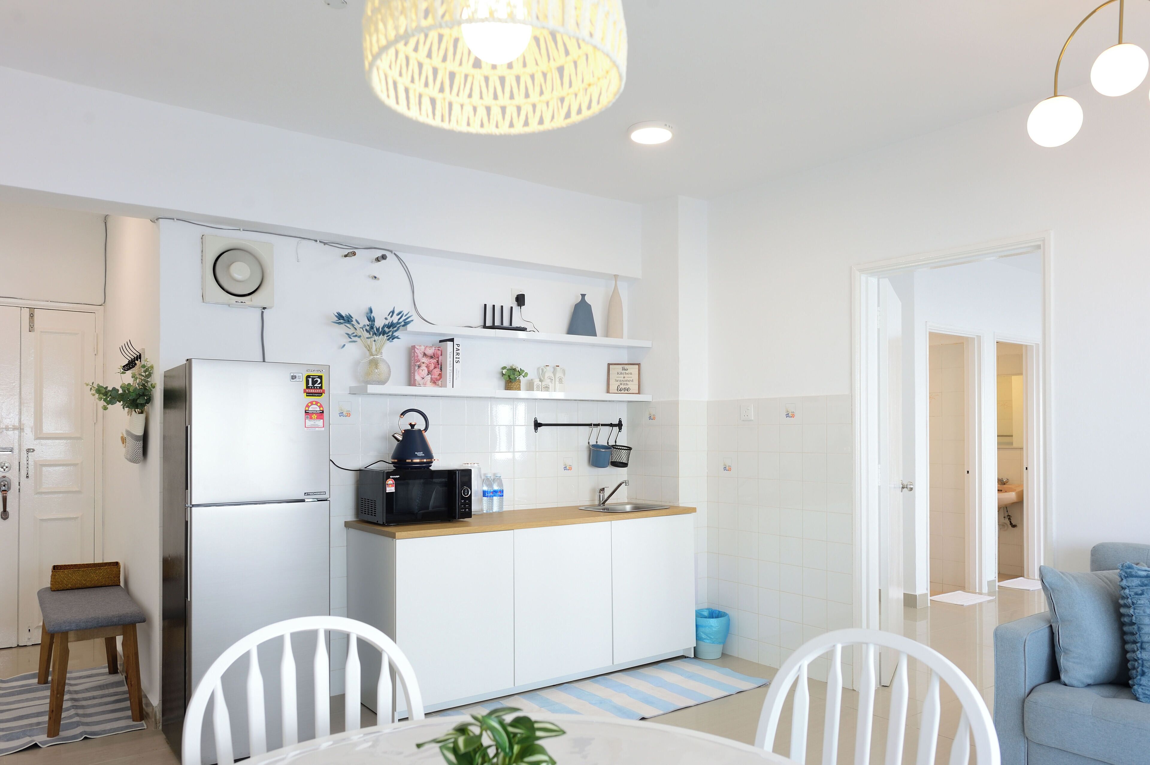 Cocina compacta con armarios blancos, un refrigerador de acero inoxidable, un microondas y un lavamanos. Cuenta con una pequeña mesa de comedor con sillas, un tapete azul y blanco a rayas, y una lámpara colgante.