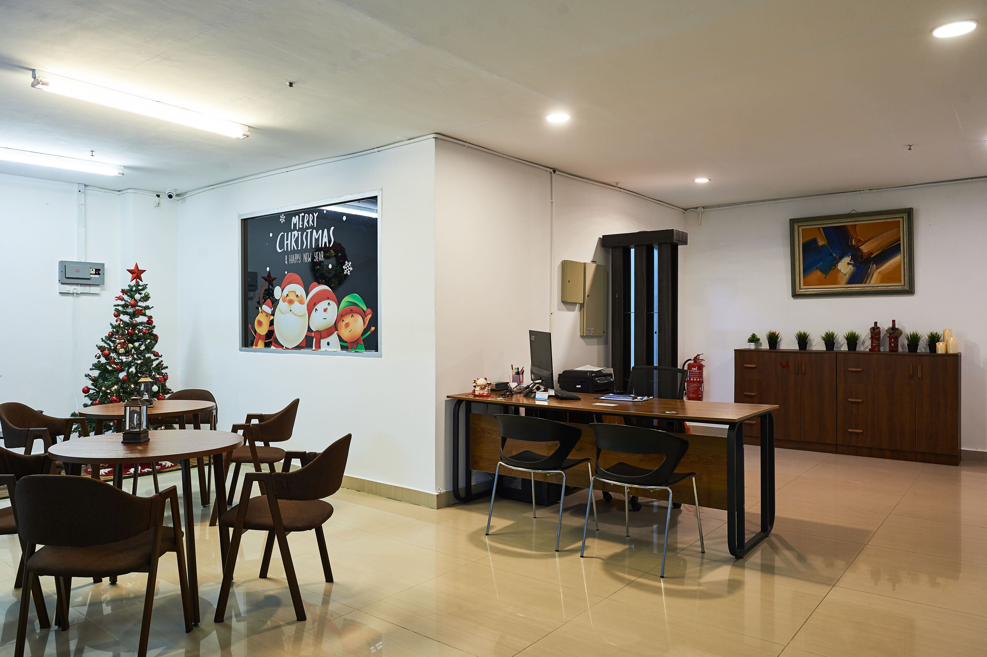A modern office space with a Christmas tree, a festive poster, a desk with a computer, and a cabinet with plants.